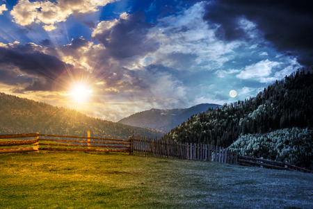 day and night time change concept. wooden fence through the grassy meadow in mountains. beautiful Carpathian countryside landscape with cloudy sky with sun and moonの写真素材