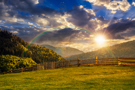 wooden fence through the grassy meadow in mountains. beautiful Carpathian countryside landscape with cloudy sky at sunset with rainbowの写真素材