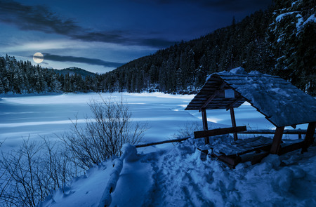 wooden bower in snowy winter spruce forest. beautiful mountainous landscape near snow covered frozen lake at night in full moon lightの写真素材
