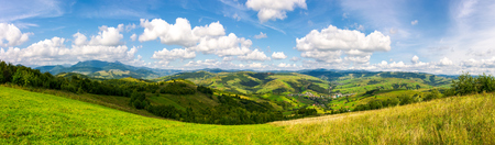 panorama of mountainous countryside. lovely countryside scenery in early autumn with grassy field on hillside, village down in the valley and clouds on a blue sky over the distant ridgeの写真素材