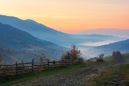 amazing beautiful rural area at dawn. morning in the carpathian mountains. fog in the distant valley. fence along the gravel roadの写真素材