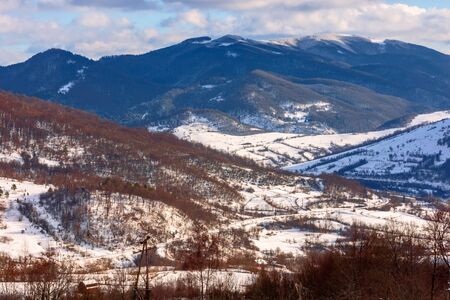 mountainous rural landscape on a sunny winter day. snow covered fields on hills. village in the distant valley. landscape with clouds on the skyの写真素材