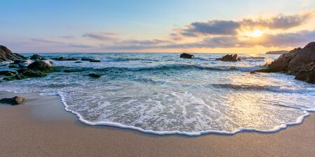 sunrise on the beach. beautiful summer scenery. rocks on the sand. calm waves on the water. clouds on the sky. wide panoramic viewの写真素材