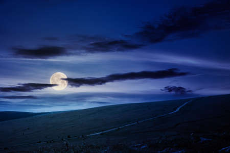 travel carpathian mountains in summer at night. road through green grassy meadows in the distance in full moon light. idyllic landscape with clouds on the blue sky.の写真素材