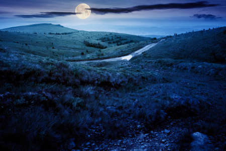 mountain road through grassy meadow at night. wonderful summer adventure in full moon light. clouds...