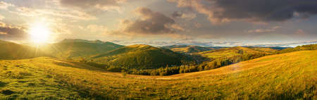 mountainous countryside landscape at sunset. panorama of a grassy rural field on the hill in evening light. village in the distant valley. clouds on the skyの写真素材