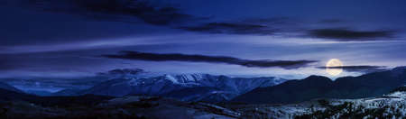 mountainous rural panorama landscape in springtime at night. beautiful scenery beneath a sky with clouds in full moon light. grass covered hill rolling in to the distant ridgeの写真素材