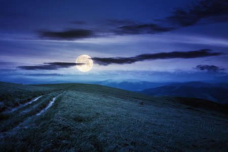 country road through alpine meadow at night. beautiful nature landscape in summer. scenery with open view in to the distant ridge and valley in full moon light. sky with clouds above the horizonの写真素材