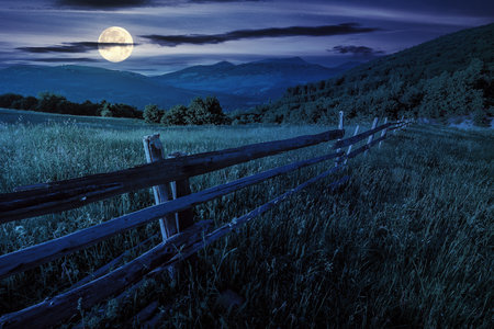 rural landscape at night. fence near the meadow and trees on the hillside. enchanted carpathian countryside scenery in full moon lightの写真素材