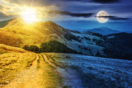 road through large meadow on the hill of mountain range with sun and moon at twilight. day and night time change concept. mysterious countryside scenery in morning lightの写真素材