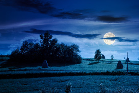 countryside landscape in summer at night. beautiful scenery with haystack on and rural field on steep green hill under blue sky with clouds in full moon lightの写真素材