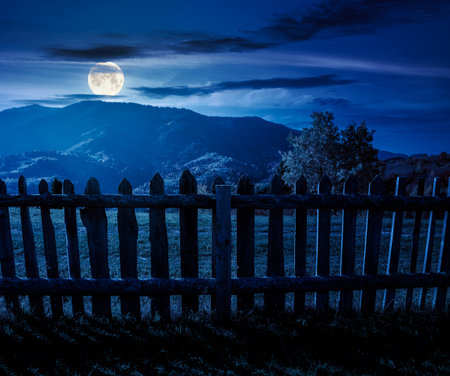 mountainous rural landscape in autumn at night. field behind the wooden fence in full moon light. scenery in fall season. forest on the hill. ridge in the distanceの写真素材