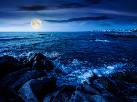 night landscape of rocky sea coast. beautiful full moon over horizon. summer coastline scenic view. composite scene during midnight. magical seascape vacations. waves and boulders in the darkの写真素材