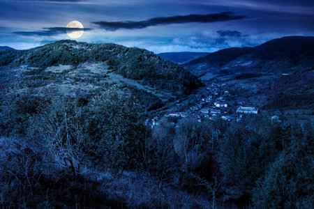 autumn landscape in mountains at night. beautiful view of countryside. colorful mountains in the background. cloudy sky. picturesque rural scenery with village in the valley in full moon lightの写真素材