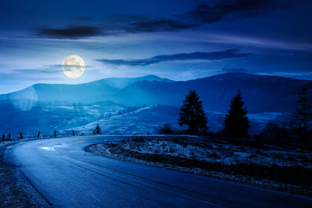 country road in mountains at night. beautiful countryside landscape in late autumn. fir trees on the roadside in full moon light. scenic view of blue sky with clouds above the distant ridge in snowの写真素材