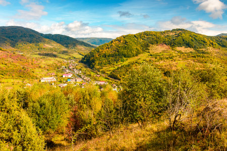 autumn landscape in mountains. beautiful view of countryside in europe. colorful carpathian alps of ukraine in the background. cloudy sky. picturesque rural scenery with village in the valleyの写真素材