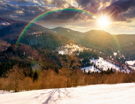 snow covered winter landscape of mountains at sunset. scenic view of countryside scenery with forested hills in evening light. fields on slopes. rural life storytelling image under the rainbowの写真素材