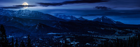 panorama of Mountain range in summer at night. village in the valley in full moon light. clouds on the sky and rolling hills. view from the topの写真素材