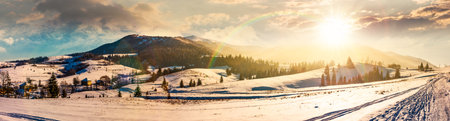 forested valley near village at sunset. landscape panorama with mountain ridge in winter in evening light. snow covered rural fields and rolling hills. cinematic image under the rainbowの写真素材