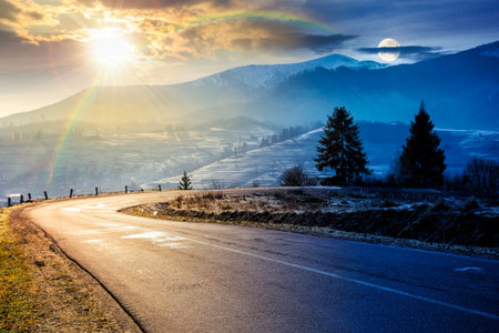 country road in mountains. day and night time change concept. beautiful countryside landscape in late autumn. fir trees on the roadside with sun and moon. scenic view of distant ridge in snowの写真素材