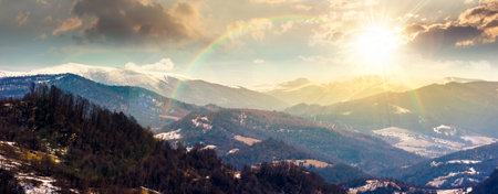 panorama of rolling hills and mountain ridge with snow cowered tops at sunset. beautiful landscape in springtime cold weather in evening light. timeless nostalgia with rainbowの写真素材