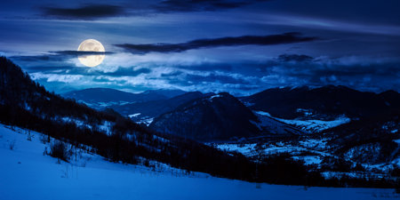 panorama of winter countryside in mountains at night. wonderful forest on the snow covered hills in full moon light. village down in the distant valley.の写真素材