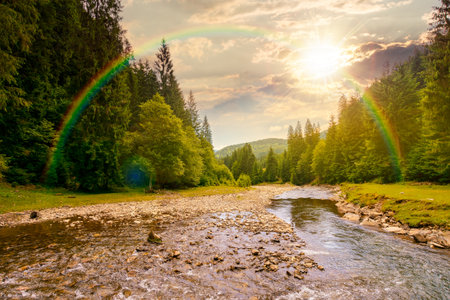 landscape with mountains, forest and a river in front at sunset. beautiful scenery in summer in evening light. water flowing through ecology and sustainability storytelling image under the rainbowの写真素材