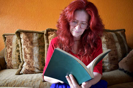Senior woman dressed in blue skirt and red blouse, sitting in a brown cloth armchair, reading or studying a green book in the living roomの写真素材