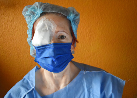 Woman operated on the right eye, wears a disposable gown and a disposable cap, brings the eye covered with a bandage, wears a blue maskの写真素材
