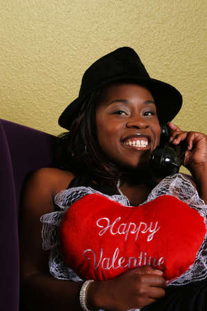 african american woman close up portrait talking on the phone with her boyfriend and holding "happy valentine" pillowの写真素材