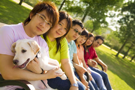 a group of asian friends sitting down on a bench outdoor with puppy labrador looking at camera with...