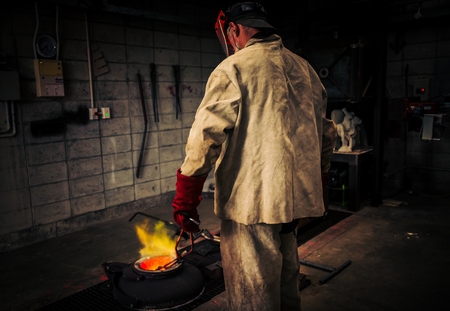 Mature man checks heat of crucible for hot metal bronze pour, using plaster moulds. An art school...