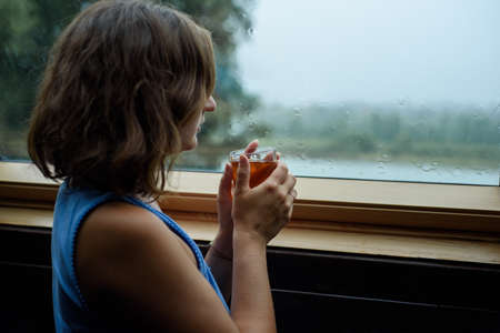Woman holding a cup of hot tea by the window in rainy weather