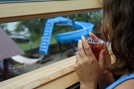 A woman drinks hot tea from a cup by the window in rainy weather against the backdrop of water...