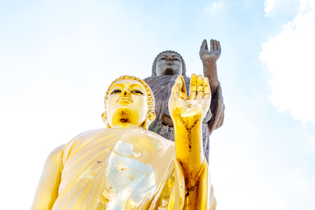 Golden Buddha statue focused on head in peace hand sign (Okay pose)  with big granite Buddha statue behind on blue sky with cloud on sunny dayの写真素材