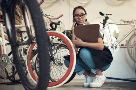 Is it the white or pink one. Full length shot of an attractive young woman crouching in her bicycle shop and using her cellphone while holding a clipboard.の写真素材
