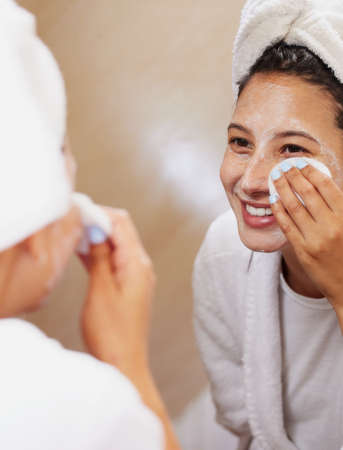 I know a few tricks when it comes to achieving flawless skin. Shot of a beautiful young woman looking in the mirror while cleaning her face.の写真素材
