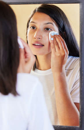 Getting cleaned up before bedtime. Shot of a beautiful young woman looking in the mirror while cleaning her face.の写真素材