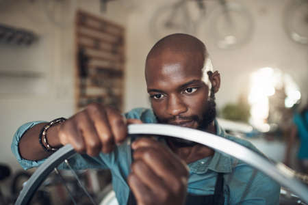 This spoke keeps popping out. Shot of a handsome young man standing alone in his shop and repairing a bicycle wheel.の写真素材