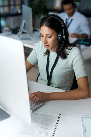 Dialled in and getting it done. Shot of a young woman using a headset and computer in a modern...