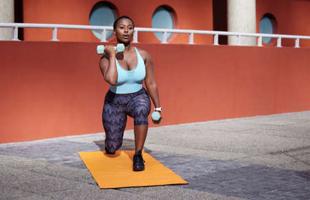 Your only worthy competitor is YOU. Shot of a young woman on a gym mat using dummbells against an...