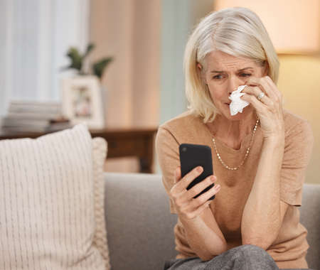 2020, the year marked by trauma. Shot of a mature woman using a smartphone and crying on the sofa at...