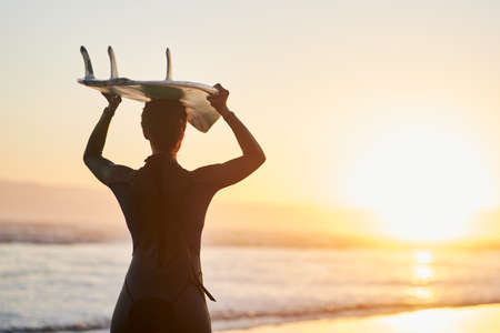 Surfing is a way of life. Rearview shot of a female surfer carrying her surfboard over her head at the beach.の写真素材