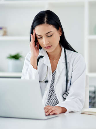 When the doctor needs a doctor. Shot of a young doctor looking stressed while using a laptop at her desk.の写真素材