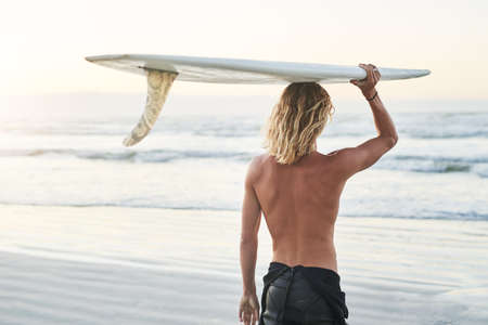 Hes riding the waves today. Rearview shot of a young surfer carrying his surfboard over his head at the beach.の写真素材