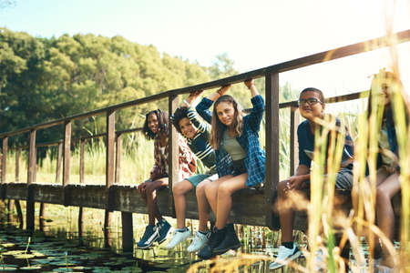 Life is sweet up the creek. Shot of a group of teenagers sitting on a bridge in nature at summer camp.の写真素材