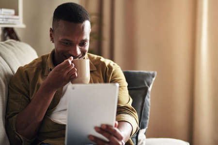 Social media is too interesting today. Cropped shot of a handsome young man drinking coffee while using a digital tablet in his living room at home.の写真素材