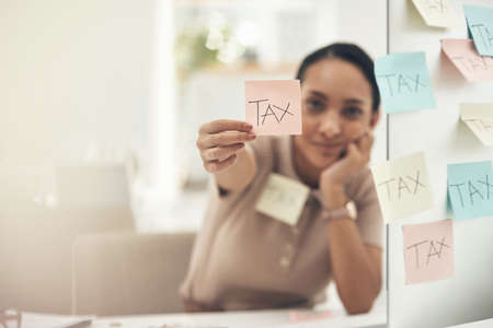 Be aware of different filing deadlines. Shot of a young businesswoman holding a note with "tax" written on it in an office.の写真素材