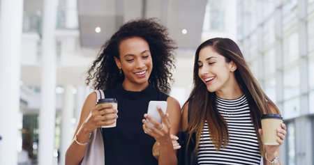Thats awesome. Cropped shot of two young businesswomen chatting and using a smartphone while walking in an office on a coffee break.の写真素材
