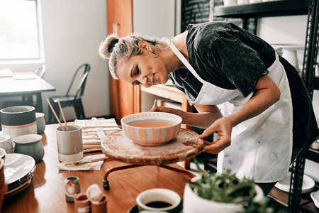 I spend time perfecting my art. Cropped shot of an attractive mature woman standing alone and painting a pottery bowl in her workshop.の写真素材